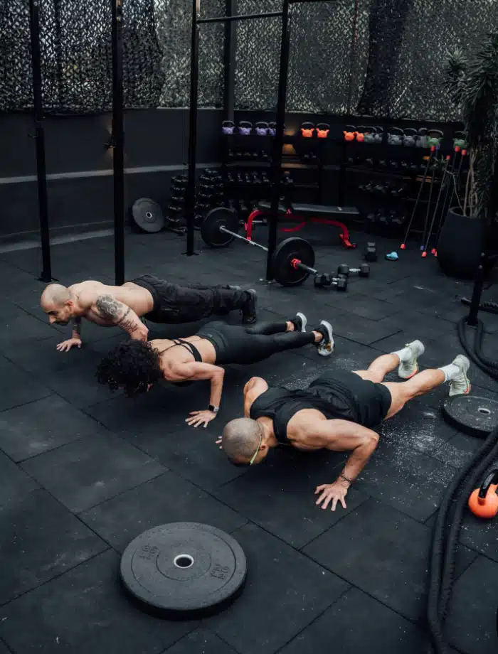 Three people are doing push-ups in a gym with black flooring. Various workout equipment, including dumbbells, kettlebells, and barbells, are visible in the background.
