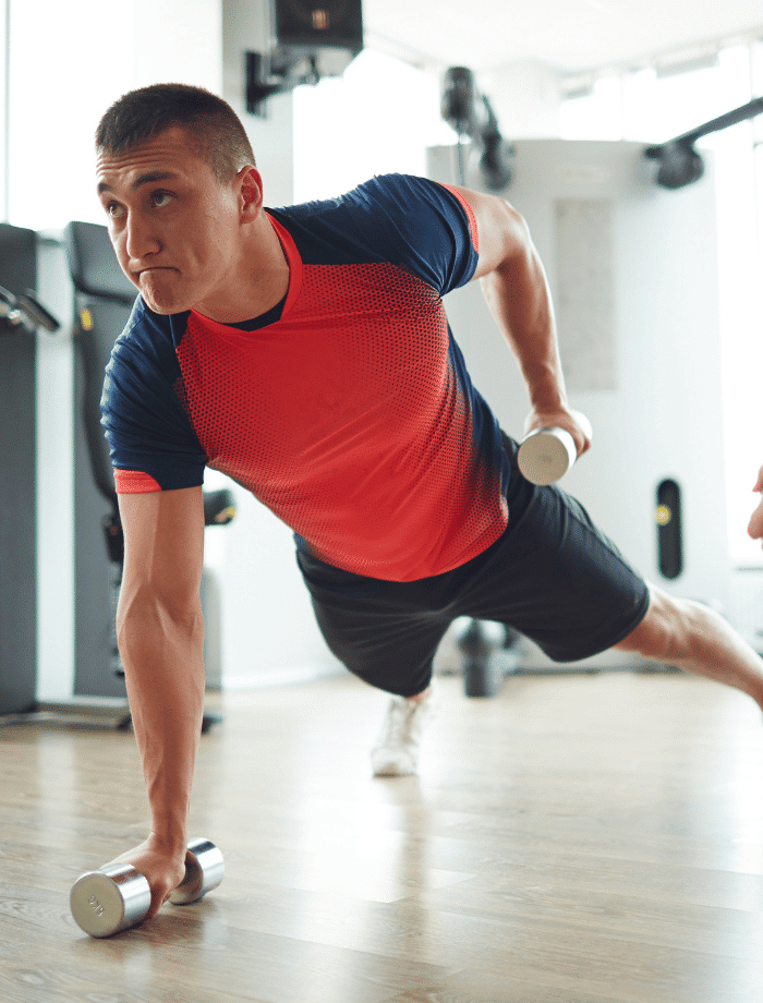 A man in a red and blue athletic shirt performs a one-arm plank row with dumbbells in a gym, focusing intensely on his workout.