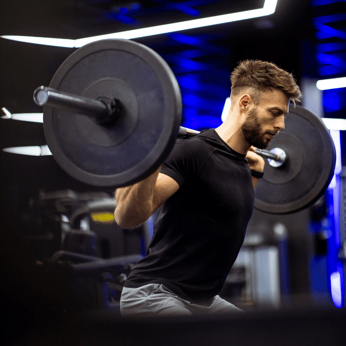 A man wearing a black t-shirt and gray shorts performs a barbell squat in a modern gym with blue lighting and fitness equipment in the background.